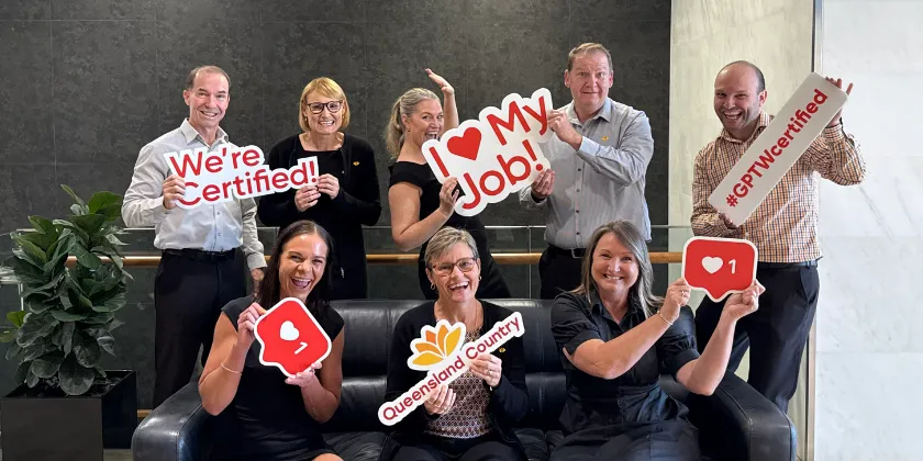 Group of Queensland Country Bank employees posing in an office lounge holding signs reading “We’re Certified,” “I ❤️ My Job!,” “#GPTWCertified,” and a Queensland Country logo, celebrating workplace certification