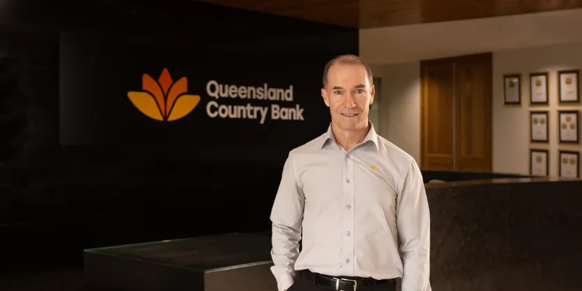 Queensland Country Bank CEO Aaron Newman standing in the lobby of the Queensland Country Centre in front of the Queensland Country Bank logo.