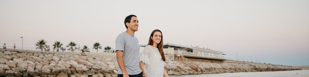 Two people standing on a rocky shoreline near the water, with a modern building and palm trees in the background under a clear sky at sunset.