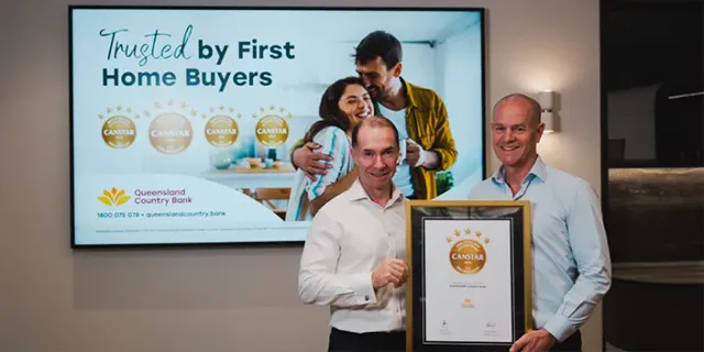 Queensland Country Bank CEO accepts a framed CANSTAR award certificate from a CANSTAR representative, standing in front of a display highlighting Queensland Country Bank's recognition as trusted by first home buyers.
