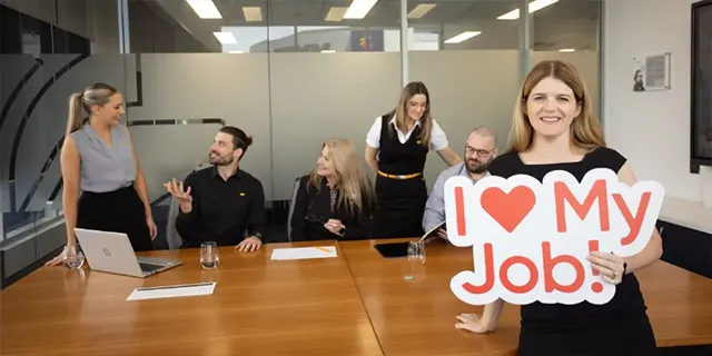 alt="Person holding an “I ♥ My Job!” sign in front of colleagues seated around a conference table in a modern office."