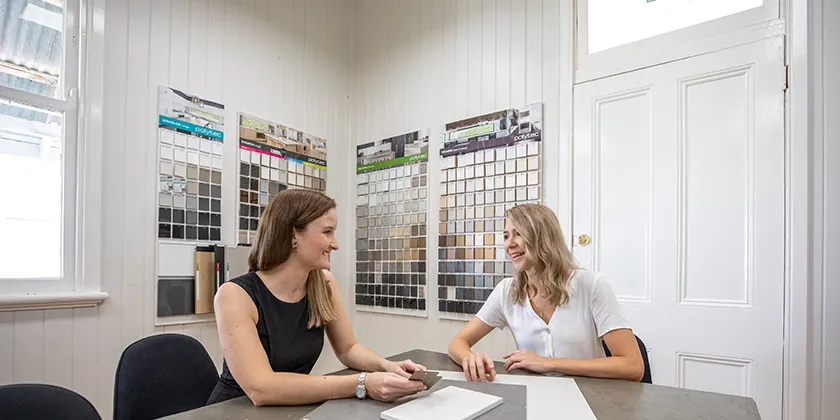 Two people seated at a table in a showroom, reviewing material samples displayed on wall boards behind them.