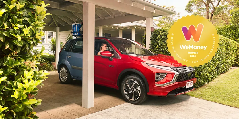 A red car parked under a carport beside a house, with a blue car positioned behind it as if transitioning into or becoming the red car to represent upgrading to a new car with a Queensland Country Bank New Car Loan. Lush hedges frame the driveway, and a yellow WeMoney 2025 award badge appears on the left side of the image.