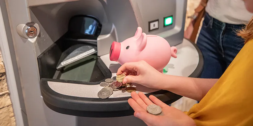 Child placing coins into a piggy bank beside an ATM, symbolising saving money.