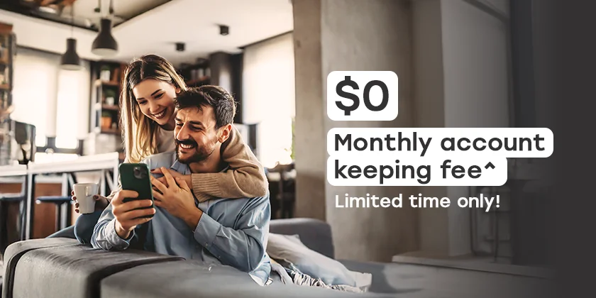 Two people sitting closely together indoors, looking at a smartphone in a cozy, modern kitchen setting, with promotional text on the right advertising $0 monthly account‑keeping fees for a limited time.