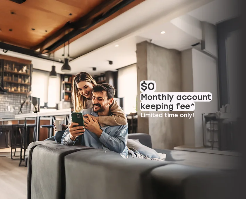 Two people sitting closely together indoors, looking at a smartphone in a cozy, modern kitchen setting, with promotional text on the right advertising $0 monthly account‑keeping fees for a limited time.
