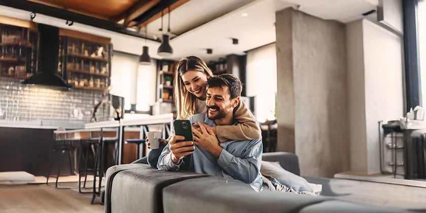 Two people sitting closely together indoors, looking at a smartphone in a cozy, modern kitchen setting.