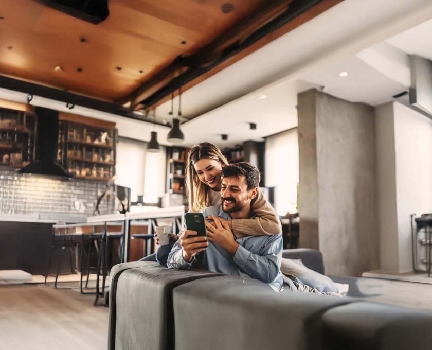Two people sitting closely together indoors, looking at a smartphone in a cozy, modern kitchen setting.
