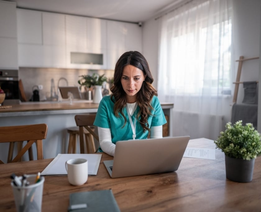 Person in medical scrubs working on a laptop at a wooden table in a bright kitchen with papers, a coffee mug, and a potted plant.